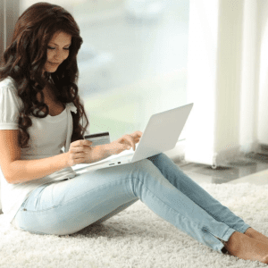 cute girl sitting by window using laptop holding credit card and smiling at e1hx5xvbx F0000